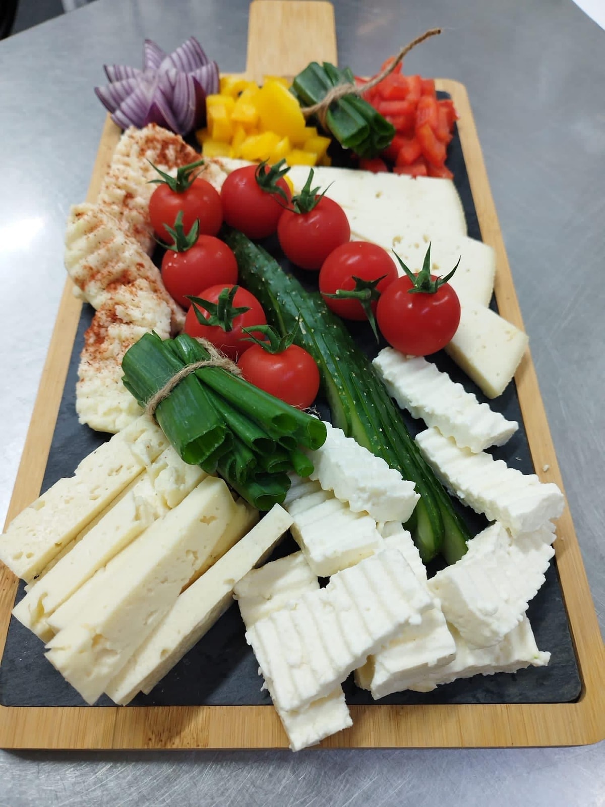 A wooden serving board is arranged with an assortment of fresh ingredients. Cherry tomatoes, cucumber slices, yellow peppers, and red onion are displayed alongside various types of cheese and a light spread. Bright colors and textures create an appealing visual presentation.