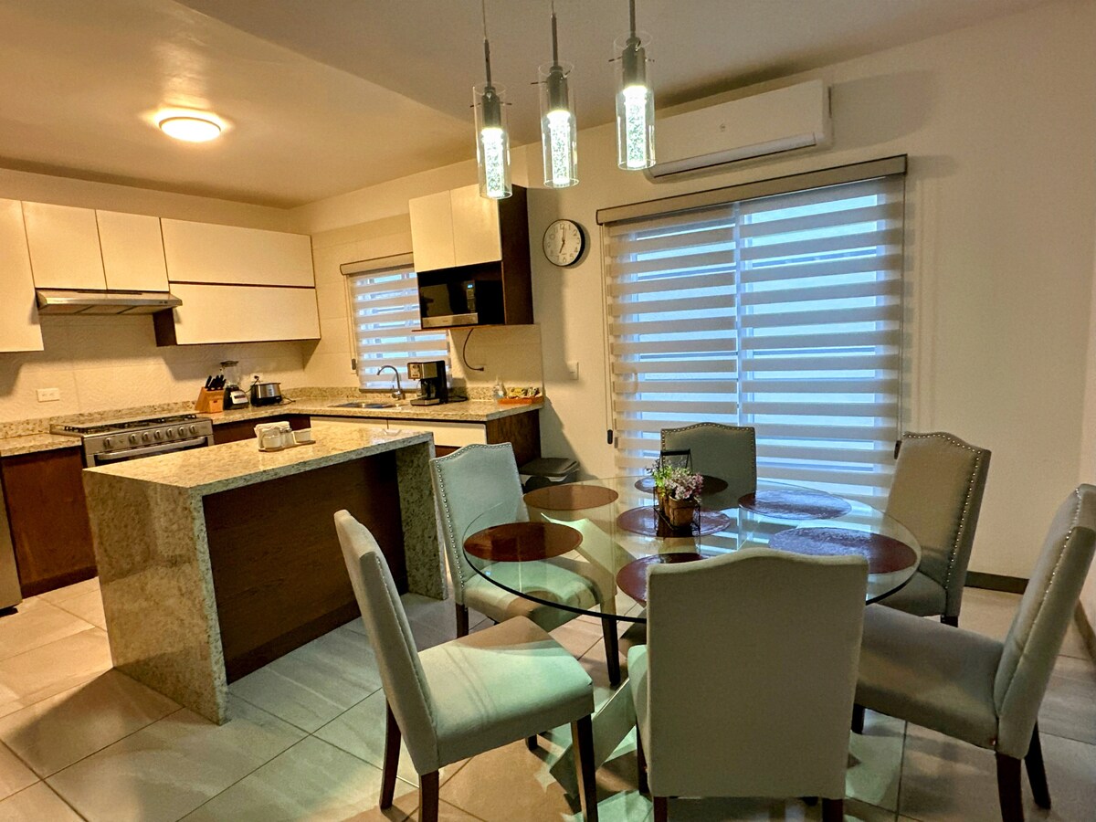 A well-equipped kitchen features modern appliances and a large island with seating. A dining area is arranged with a round glass table surrounded by six upholstered chairs. Natural light filters through blinds on two windows, providing a bright and airy atmosphere.