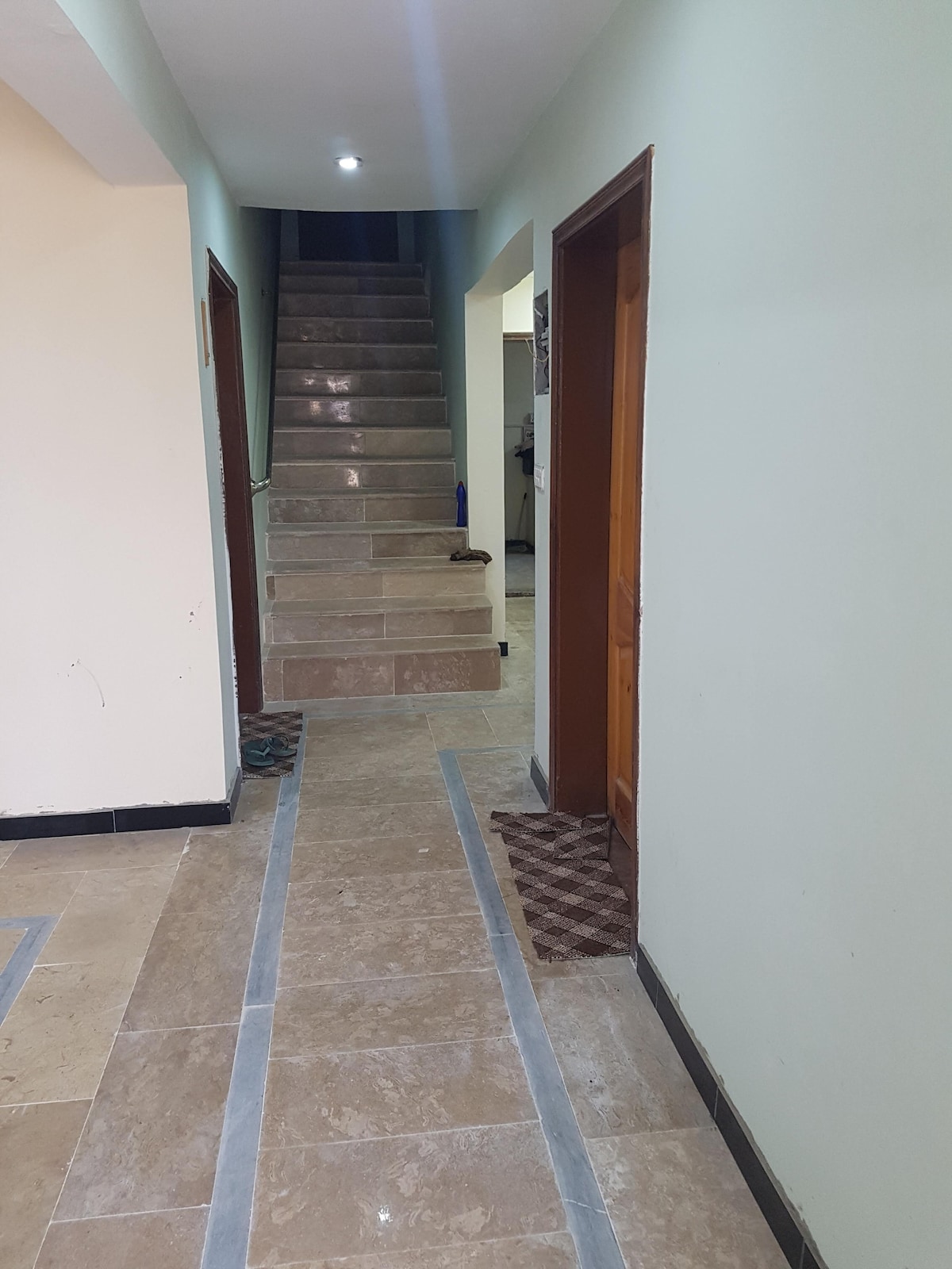 A well-lit hallway is seen, featuring smooth stone flooring with beige and gray tones. Doors to rooms are located on either side, while a staircase leads to the upper floor in the background. Light fixtures provide soft illumination along the pathway.