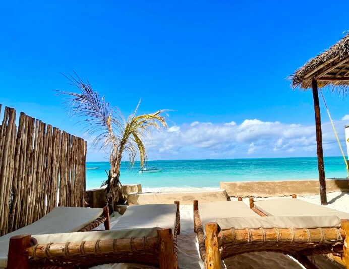 Lounge chairs are positioned on the sandy beach, inviting relaxation. A palm tree leans slightly, providing gentle shade. The turquoise Indian Ocean stretches in the background under a clear blue sky, enhancing the serene coastal atmosphere. Thoughtful beach structures frame the scene.