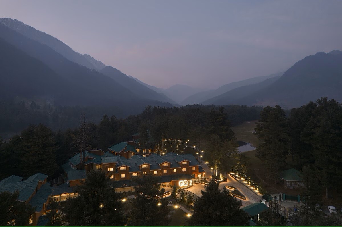 An aerial view captures a charming lodge nestled in a valley, surrounded by mountains. Soft lighting outlines the building and pathways, providing a gentle glow amidst the twilight. Lush trees and an expansive landscape enhance the tranquil setting.