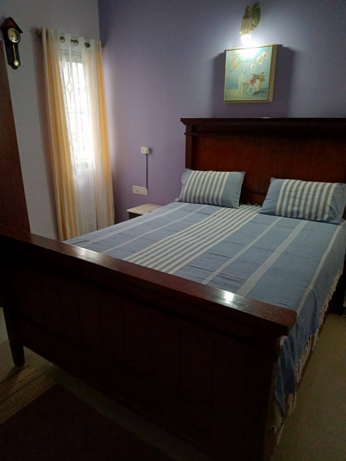 A warm-toned bedroom is presented, featuring a wooden bed adorned with a light blue striped blanket and two matching pillows. Pale purple walls provide a soft backdrop, while a framed artwork adds a touch of charm. Soft light is emitted from a wall lamp near the bed.