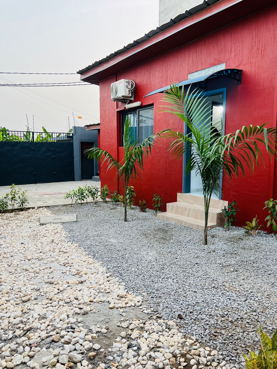 A vibrant red exterior wall contrasts with a light-colored entryway, framed by two small palm trees. A pathway leads to the door, while gravel and small plants are arranged in the surrounding garden area, enhancing the overall outdoor appearance.