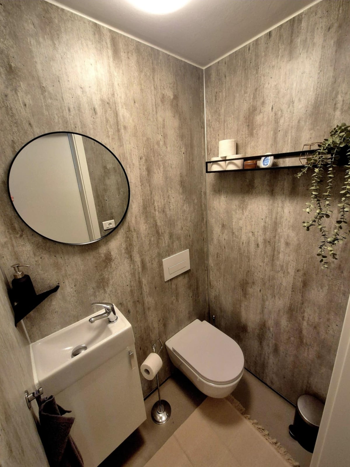 A modern bathroom features concrete-textured walls with a circular mirror above a compact sink. A wall shelf holds decorative items, while a toilet is positioned nearby. Neutral tones and simple design elements create a clean and functional space.