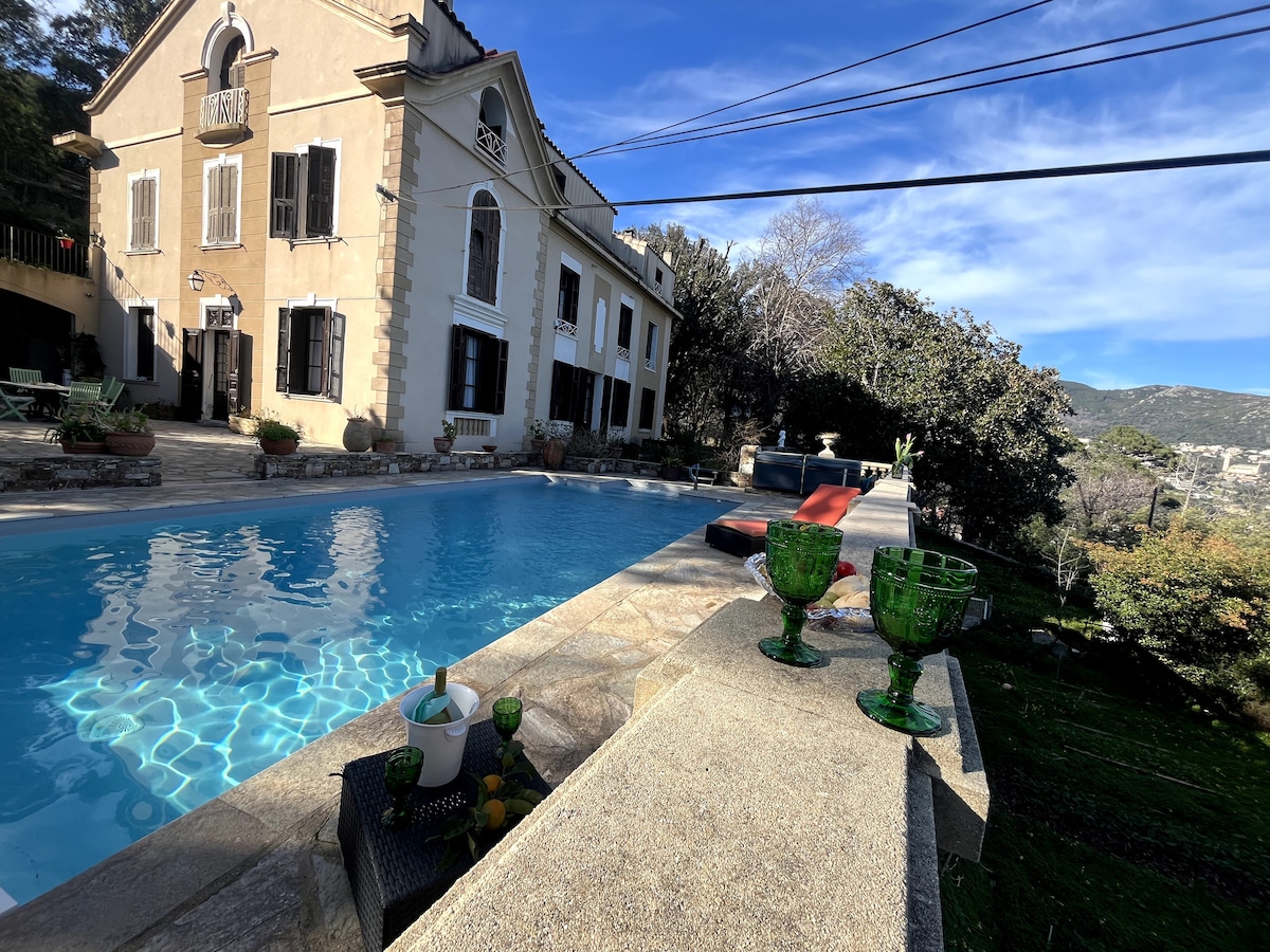 An outdoor view features a large swimming pool reflecting clear blue skies. Sunshine glistens on the water's surface. In the foreground, green glass cups rest on a stone ledge, while an inviting lounging area and a jacuzzi are visible in the background.