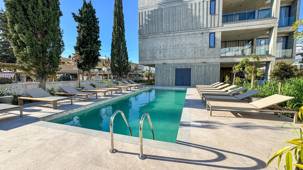 A communal pool is surrounded by lounge chairs and greenery, providing a serene outdoor setting. Modern building lines and tall trees are visible in the background. The pool features a clear, inviting water surface, and easy access steps lead into the water.