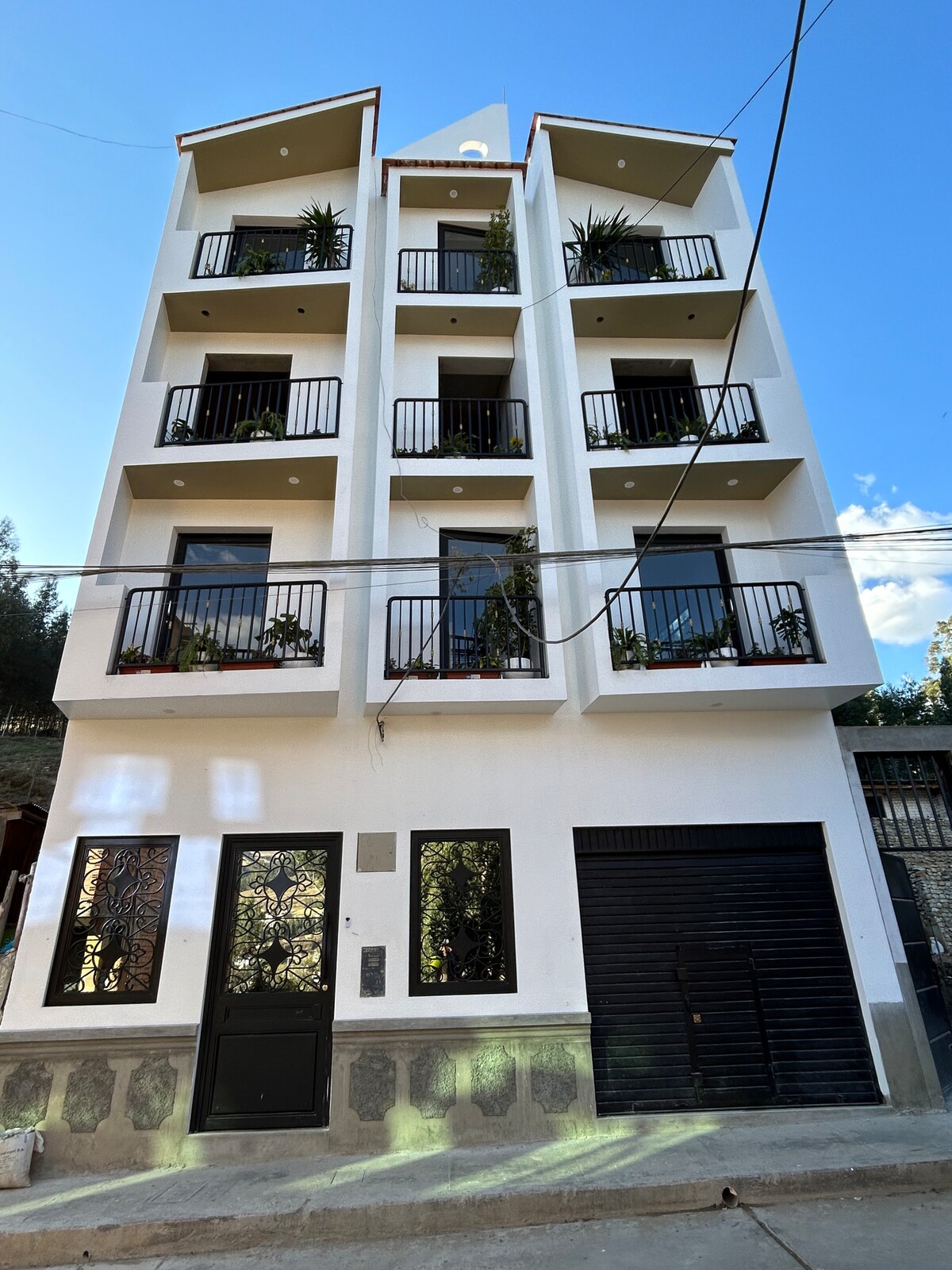 The modern multi-story structure features a clean white facade with black balconies adorned with greenery. Large windows provide ample natural light, while the entrance is marked by an imposing black door. The surrounding area reflects a tranquil environment, complemented by the building's neat design.