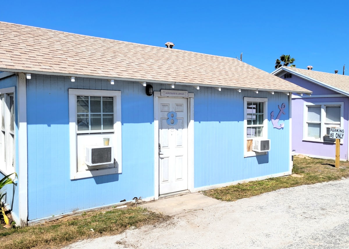 A light blue cottage with a beige roof is depicted, featuring large windows and two air conditioning units. The front door is centrally positioned with the number 8 displayed above it. Nearby, a sign indicating 'Parking Only' is visible, set against a clear blue sky.