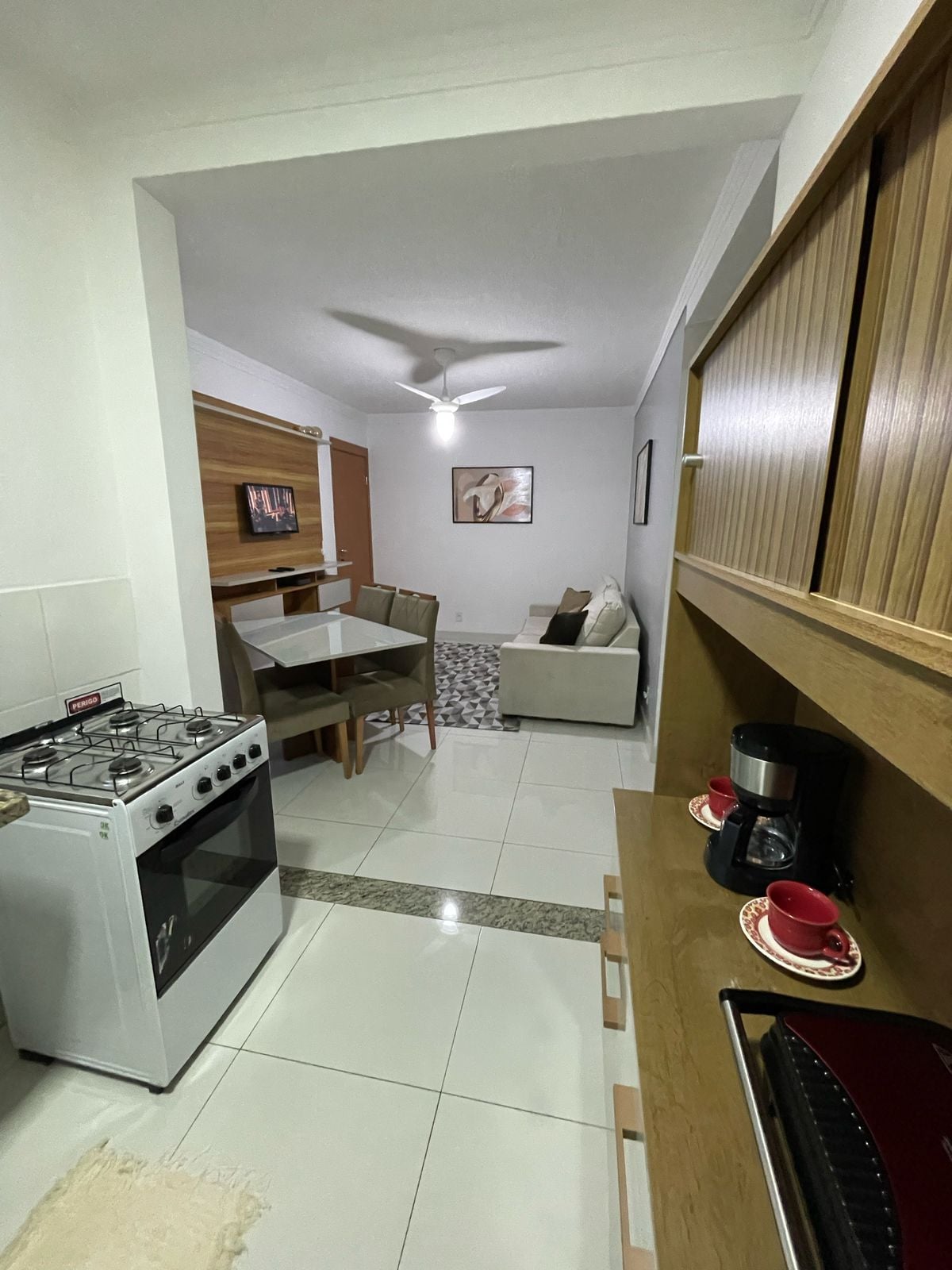A modern kitchen is seen leading into a comfortable living area. A small dining table is positioned adjacent to a sofa, with a ceiling fan overhead. The space features white tile flooring and natural wood accents, enhancing a functional and inviting environment.