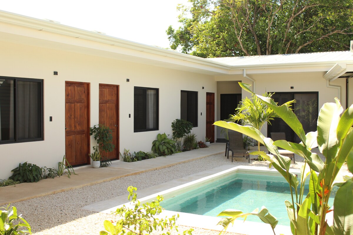 A serene courtyard features a tranquil pool surrounded by greenery. Simple wooden doors lead to individual rooms, while potted plants enhance the natural beauty. A comfortable chair sits nearby, inviting relaxation among the peaceful outdoor setting.