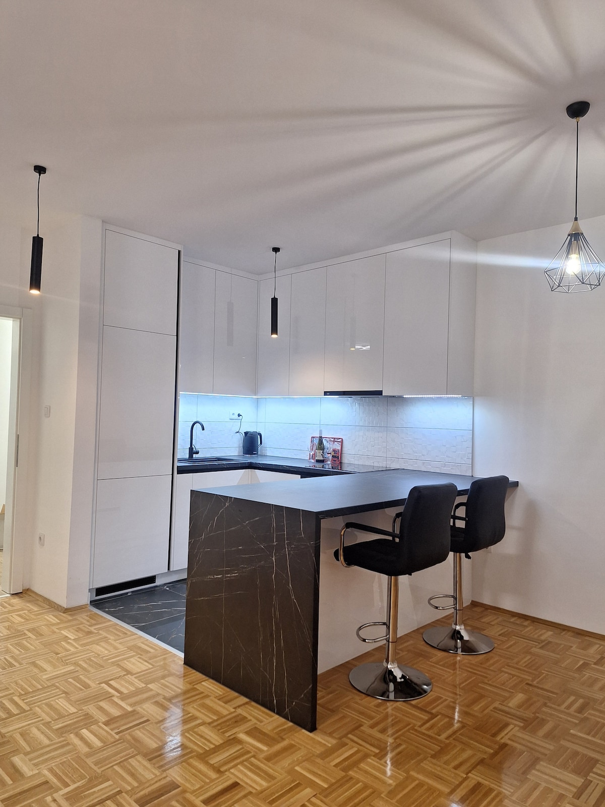 The modern kitchen is characterized by sleek white cabinetry and a contrasting dark island featuring two tall stools. Under-cabinet lighting illuminates the workspace, while pendant lights add a stylish touch to the ceiling. A patterned wooden floor completes the inviting design.