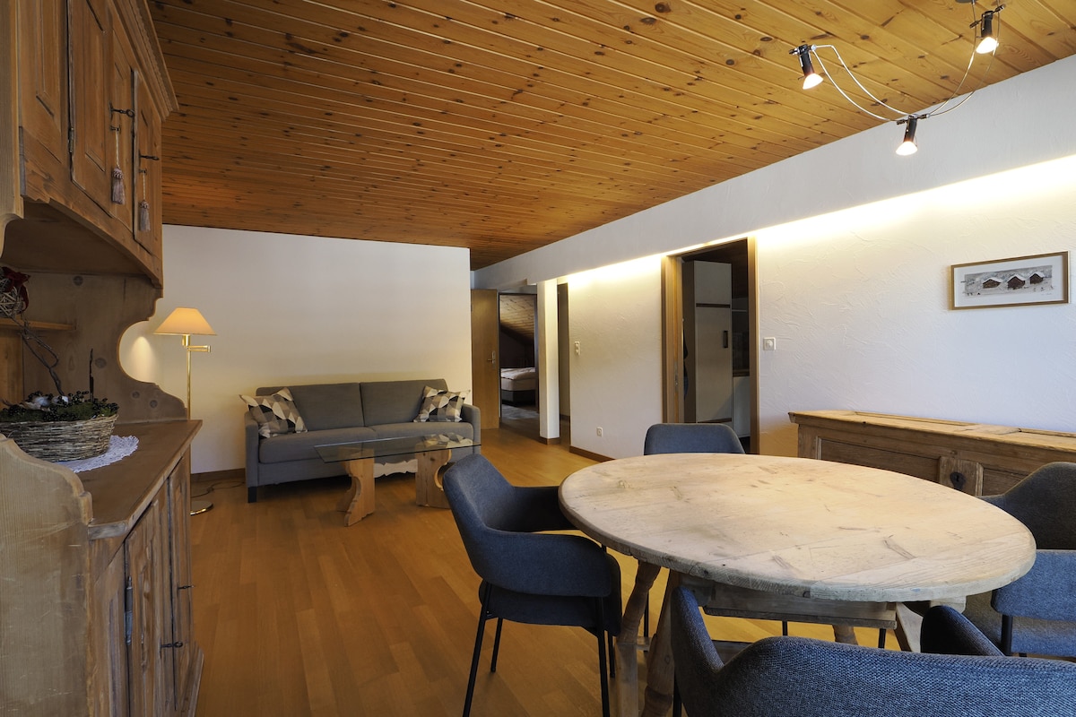 A spacious living area features wooden paneling and a high ceiling with warm lighting. A round wooden table is surrounded by blue upholstered chairs. A cozy couch sits against the wall, and a side lamp adds soft illumination. Two doors lead to additional rooms.