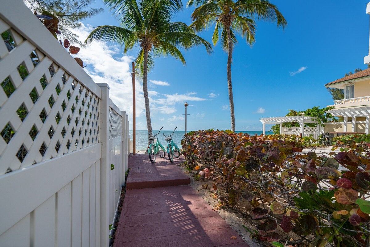 A pathway lined with vibrant greenery leads towards the beach, where shallow turquoise waters meet the shore. Two bicycles are positioned near the fence, offering guests easy access to the seaside. A clear sky with a few clouds adds to the serene coastal ambiance.