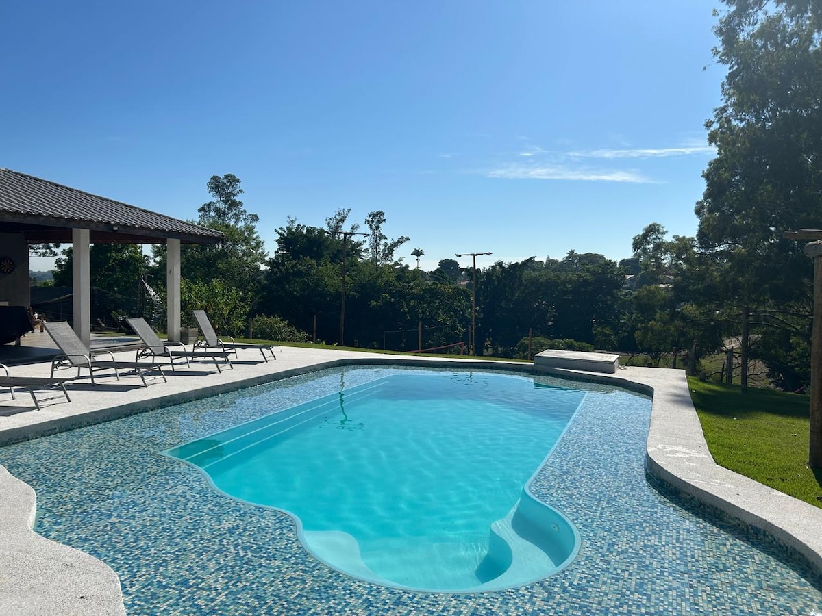 A serene pool area is visible, featuring a shaped pool with clear blue water and a decorative tile border. Lounge chairs are positioned around the pool deck, and lush greenery is seen in the background under a clear blue sky.