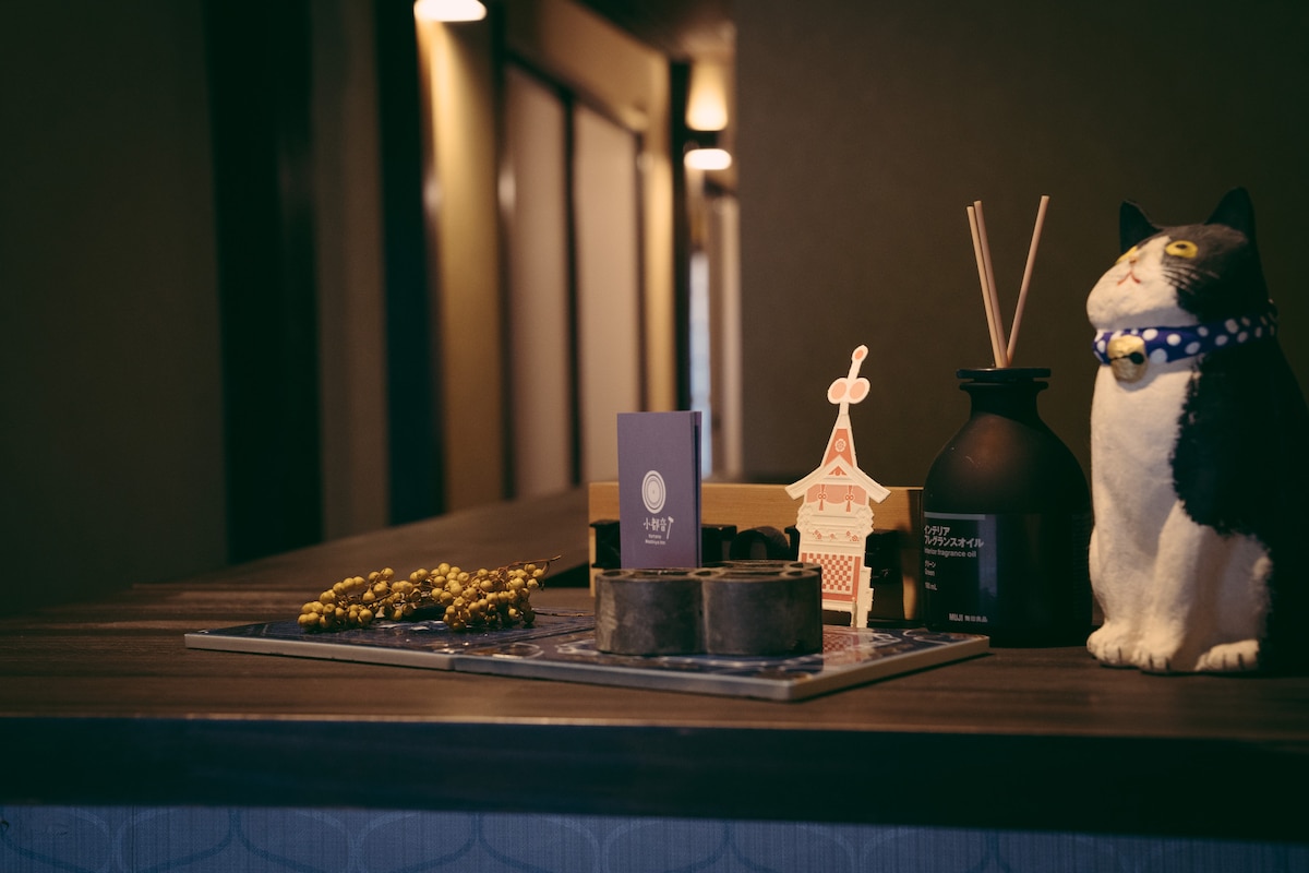 A decorative counter is featured, showcasing a variety of items. A cat figurine in a collar sits beside an arrangement of incense and decorative pieces. Dark wooden tones and soft lighting create a calm setting, inviting guests into the space.
