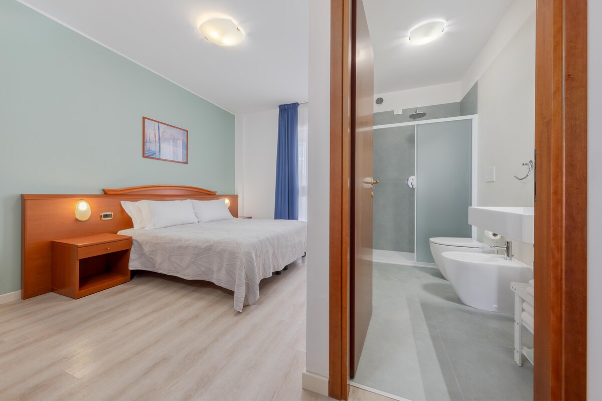 A well-lit bedroom is showcased with a spacious bed framed by light-colored walls. A bedside table sits beside the bed. An open door reveals a modern bathroom with a shower, sink, and toilet, featuring a clean and minimalist design.