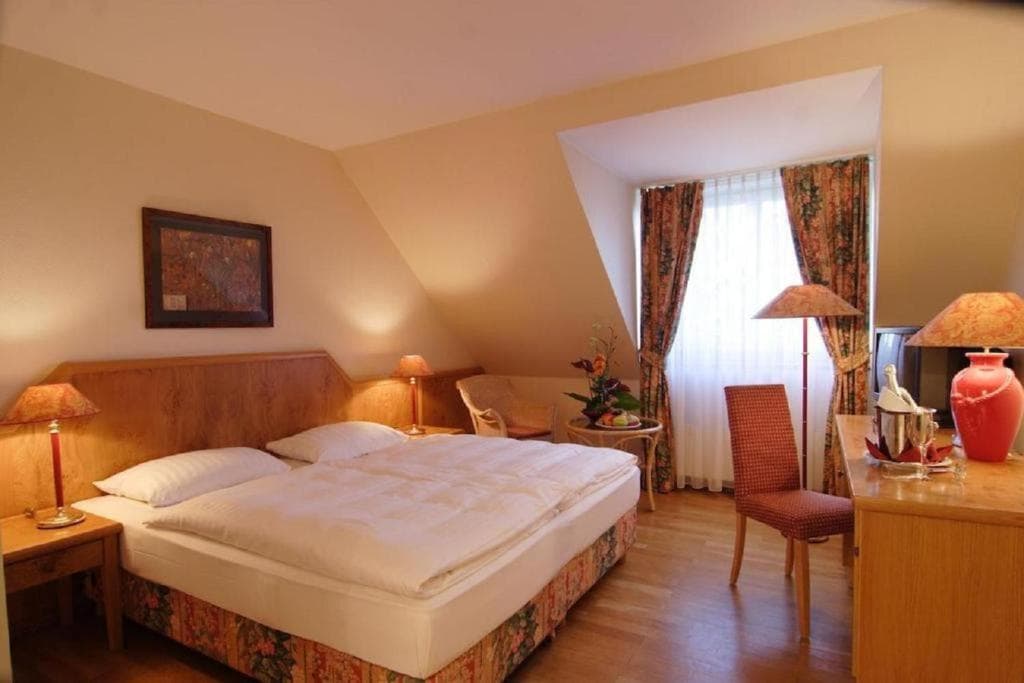 A cozy room features a spacious bed with a light quilt, surrounded by soft illumination from bedside lamps. A large window allows natural light to fill the space, while a tasteful arrangement of furniture includes a wicker chair and a wooden desk.