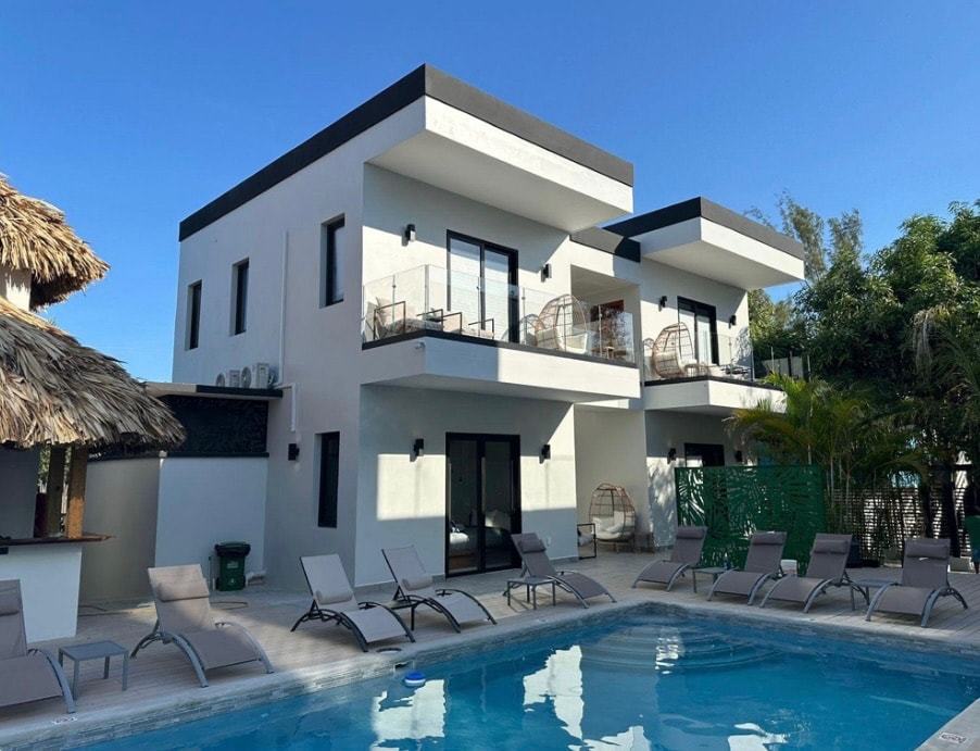 A modern two-story villa is depicted, featuring a clean, minimalist design with large glass doors. Surrounding the inviting pool, several lounge chairs are arranged, and balconies with seating provide spaces for relaxation. Lush greenery adds a natural touch to the tranquil outdoor setting.