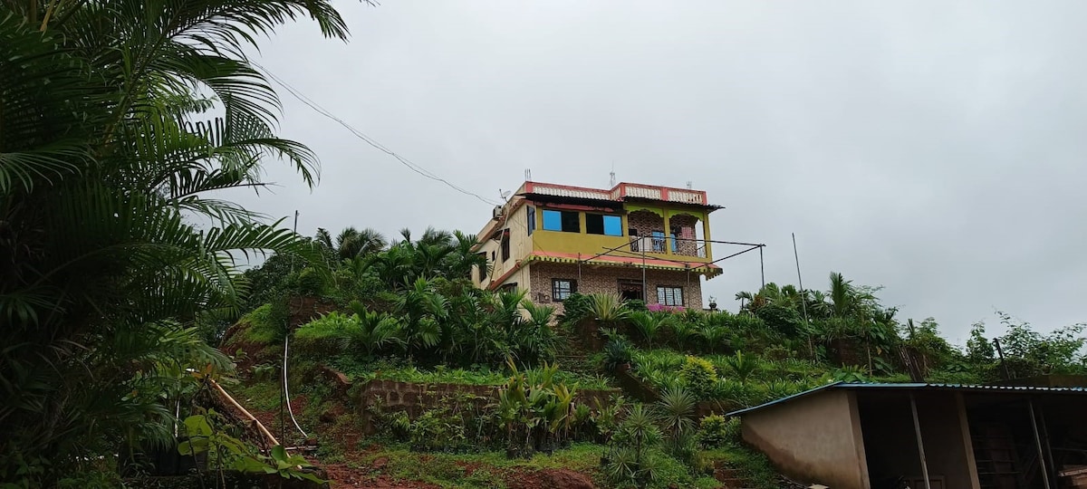 A two-story house is situated on a hillside, surrounded by lush greenery and tropical plants. The building features colorful accents, including blue and yellow accents on its exterior. A cloudy sky looms overhead, while terraced landscaping enhances the natural setting.