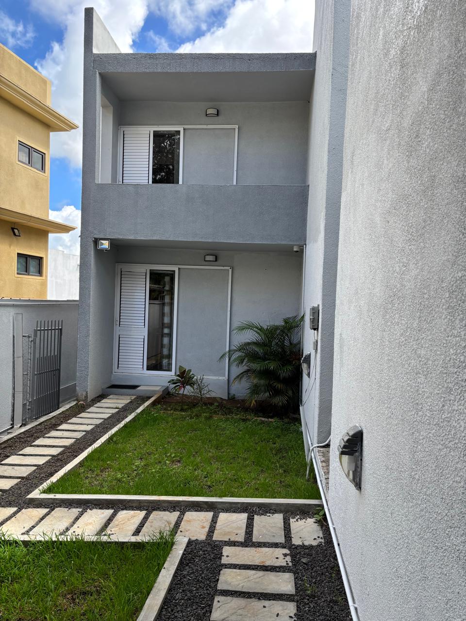 A well-maintained pathway leads through a grassy area, flanked by light-colored stone pavers. The exterior of a two-story structure is visible, featuring large windows with white shutters. Lush greenery is present beside the path, enhancing the tranquil ambiance.
