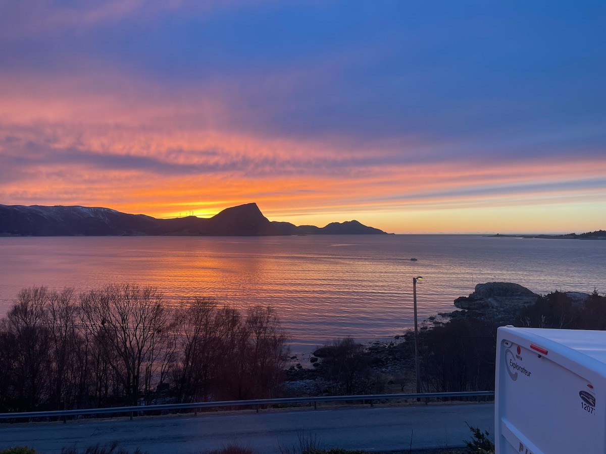 A serene view of a sunset over the water, with vibrant hues of orange, pink, and purple reflecting off the calm surface. Silhouetted mountains and trees frame the scene, creating a peaceful backdrop against the horizon.