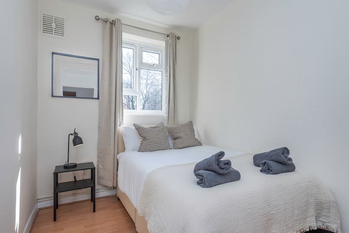 Petit lit double confortable avec draps blancs doux et oreillers moelleux. Une fenêtre avec des rideaux légers remplit la pièce de lumière naturelle. Une lampe noire élégante se trouve sur la table de chevet. Un miroir minimaliste ajoute une touche de modernité. Les serviettes grises propres sont soigneusement pliées