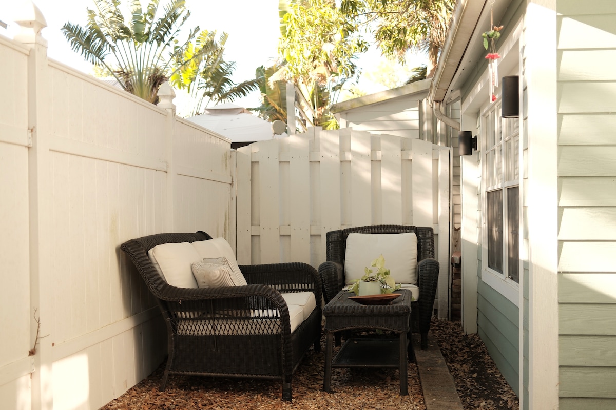 A cozy outdoor seating area is surrounded by white fencing and features two wicker chairs with soft cushions. A small table holds a potted plant, and sunlight filters through, creating a warm ambiance.