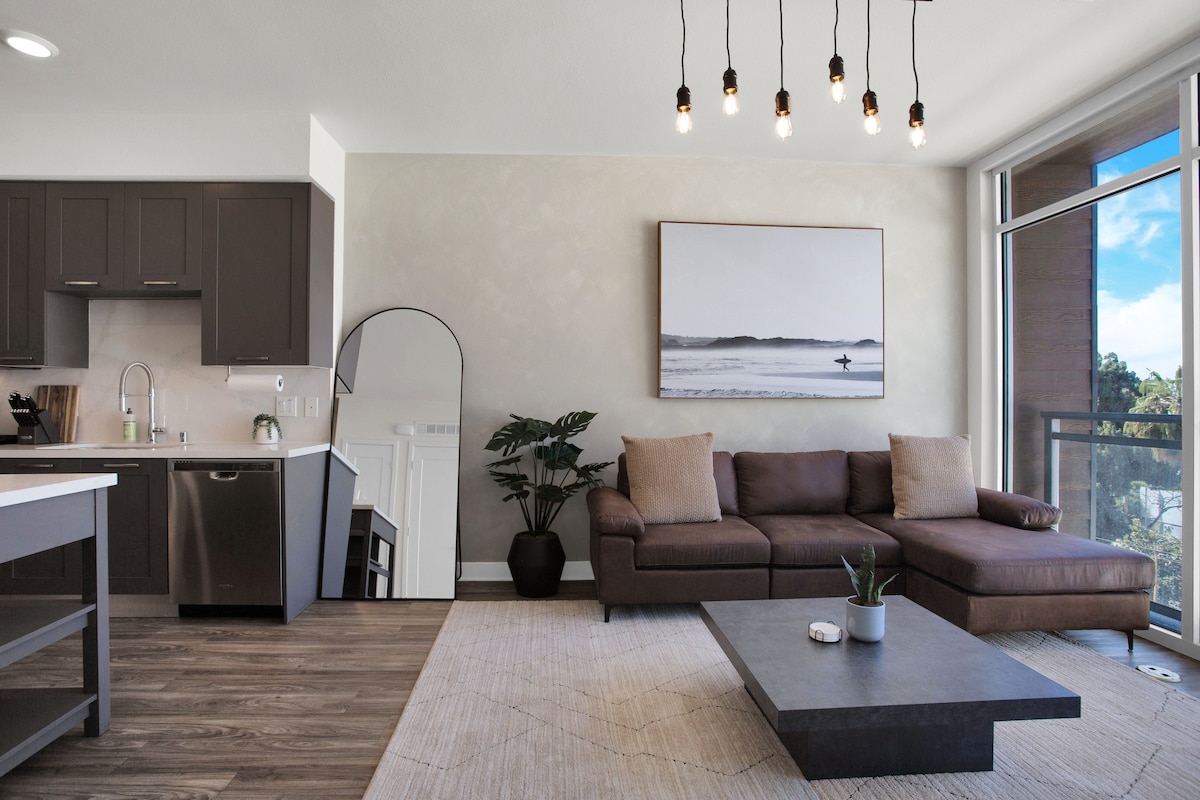 A modern living area features an L-shaped brown sofa and a rectangular coffee table positioned on a light-colored area rug. A large mirror and kitchen appliances are visible, while natural light enters through the expansive window, highlighting the neutral color palette.