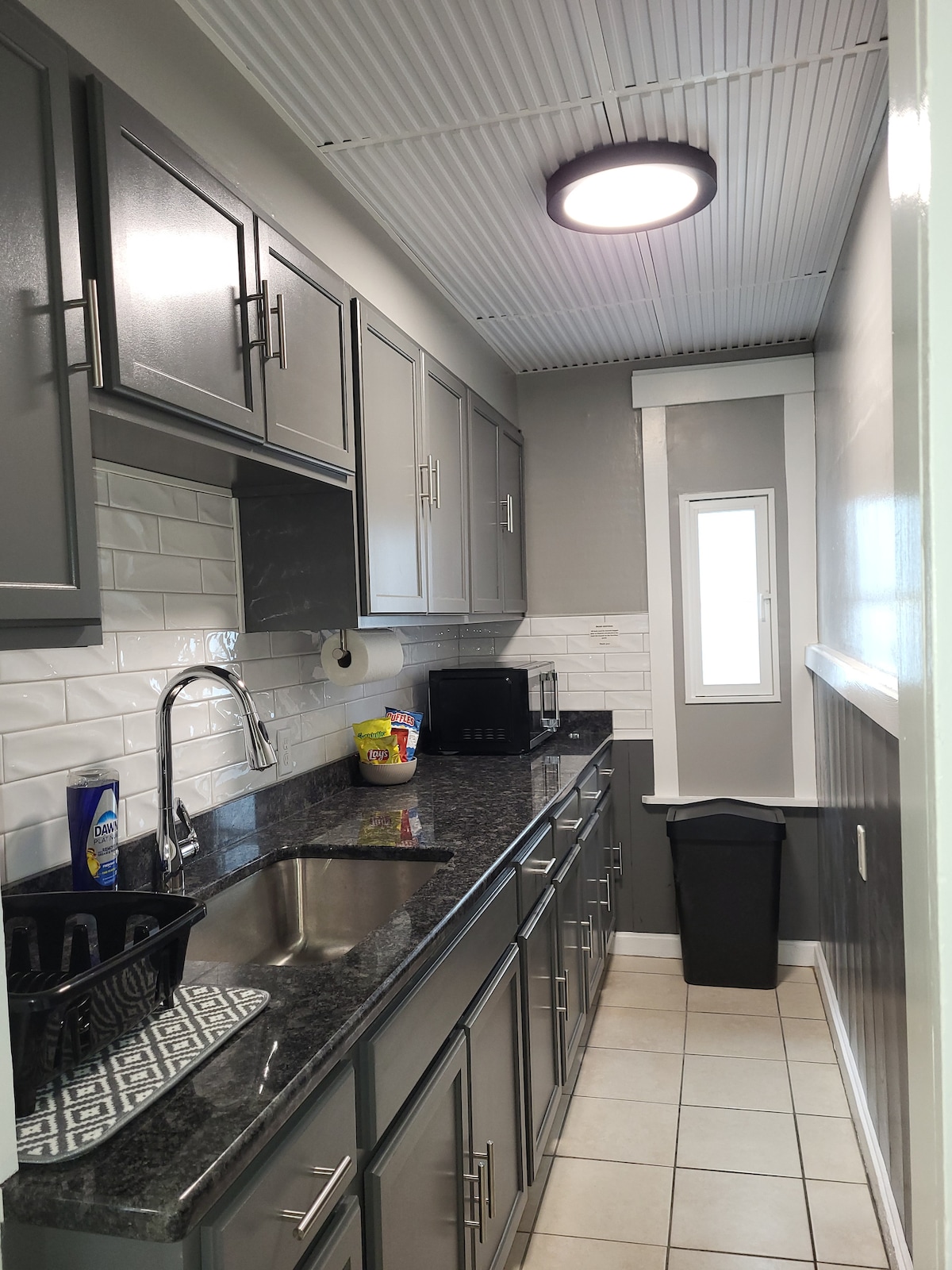 A galley kitchen is displayed with modern gray cabinetry and a sleek black countertop. The kitchen is equipped with essential appliances, including a microwave and a sink. White subway tile backsplash contrasts with the cabinetry, while a small window allows natural light into the space.