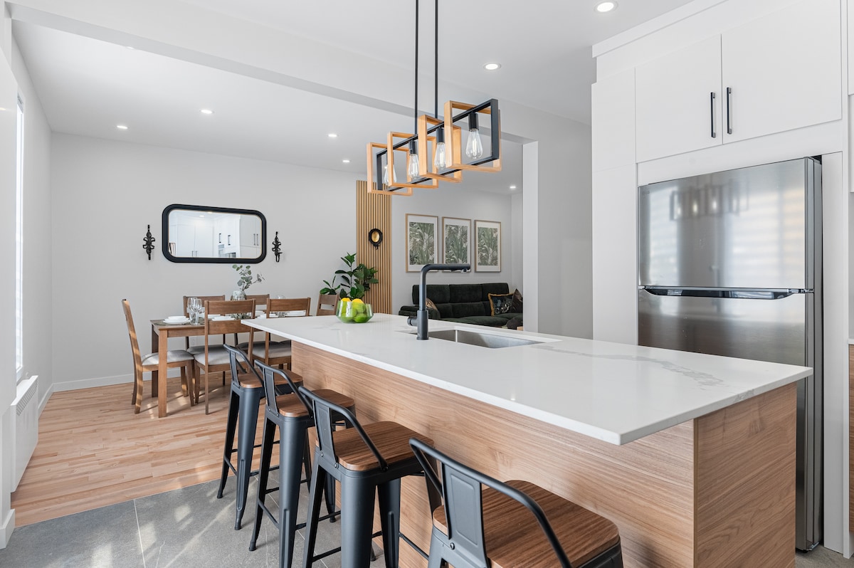 The modern kitchen features a large island with bar seating for four, complemented by a sleek light fixture above. The stainless steel appliances, including a refrigerator, are situated next to a cozy dining area visible through a decorative mirror.