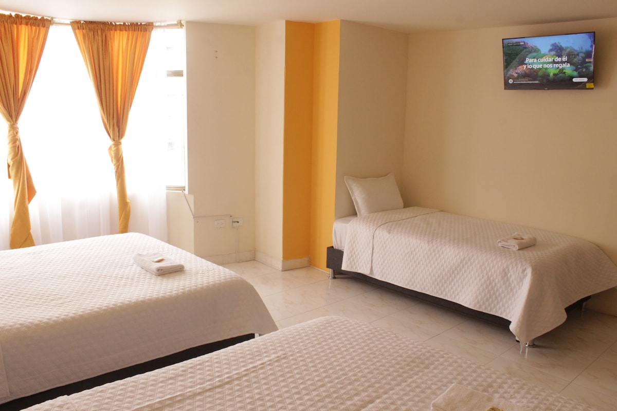 A well-appointed guest room features three spacious beds, each adorned with white bedding. Natural light is welcomed through large windows covered by sheer curtains, complemented by warm-toned drapes. A flat-screen television is mounted on the wall, providing entertainment options.