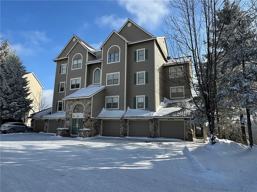A multi-story townhome is surrounded by snow-covered trees. Its exterior features a combination of stone and siding, with large windows and a welcoming entrance. A clear path leads through the snow to the garage doors, which provide access to heated parking.