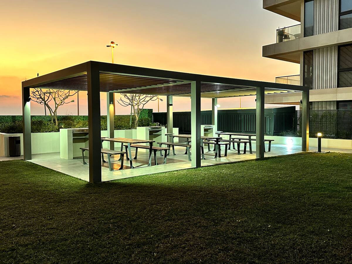 An outdoor seating area is highlighted by several picnic-style tables beneath a shaded pavilion. The space is illuminated with soft lighting, providing a welcoming ambiance against a sunset backdrop. Lush grass surrounds the area, enhancing the serene outdoor setting.