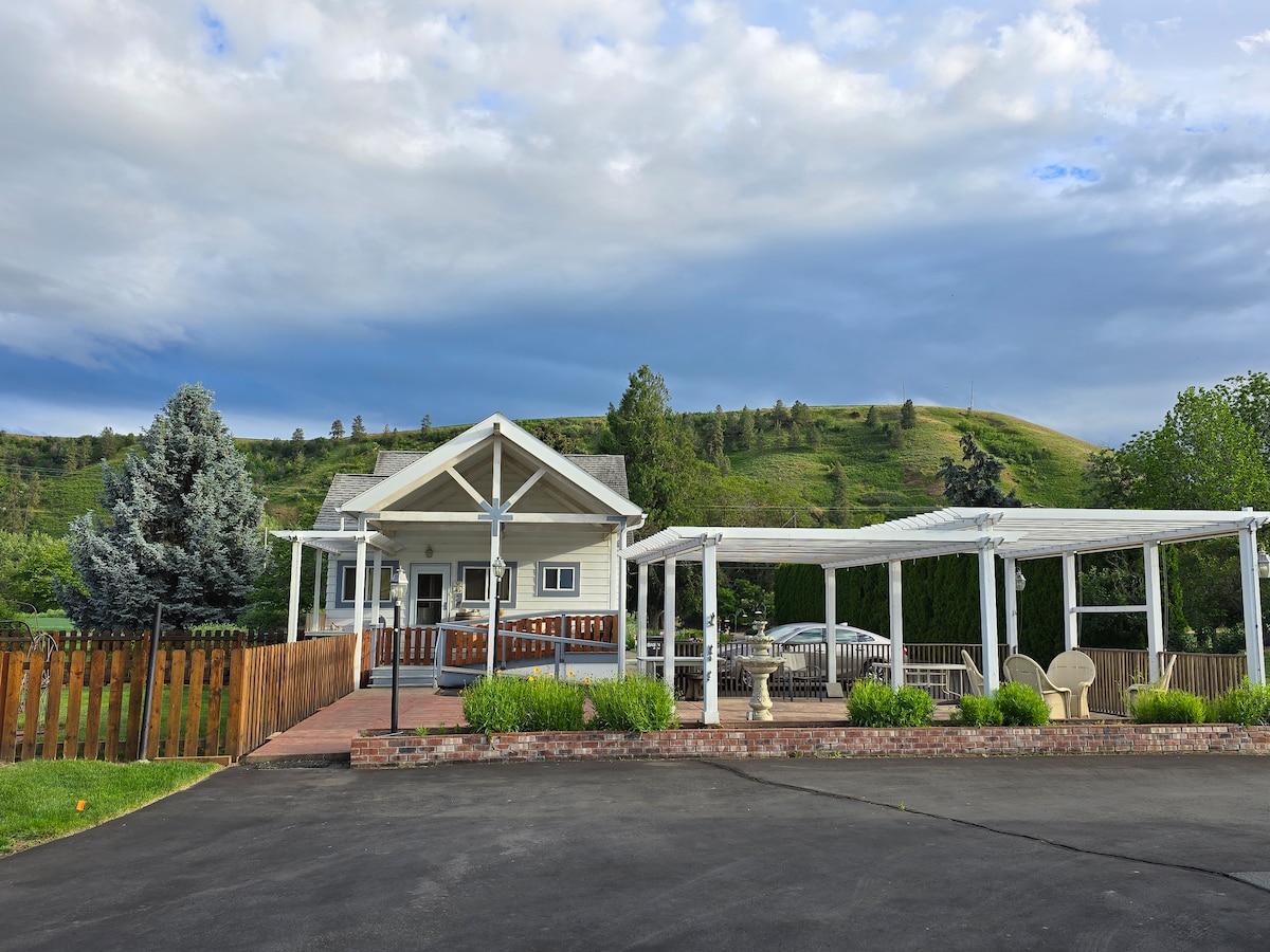 The exterior view of a charming, two-story home is displayed, featuring a welcoming front porch and a large covered patio area. Surrounding greenery and a hill in the background enhance the peaceful setting. A paved driveway leads to the entrance, bordered by neatly maintained landscaping.