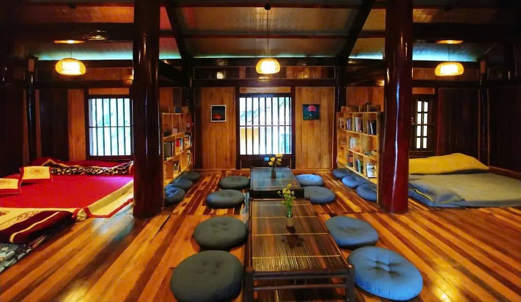 The communal space features wooden beams and flooring, showcasing an open layout with seating cushions and low tables. Bookshelves are filled with books, while natural light enters through multiple windows. Colorful blankets are arranged on the floor, inviting relaxation and interaction among guests.