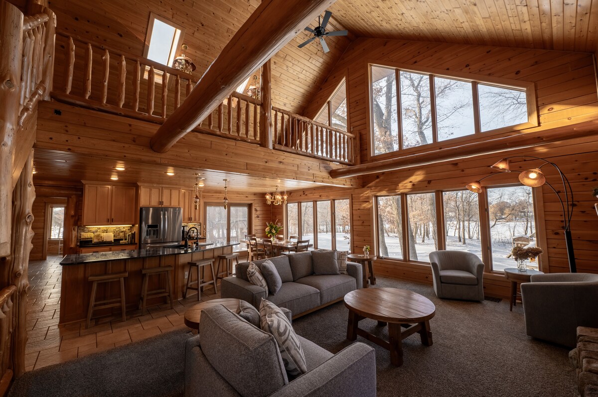 A spacious living area is highlighted by natural wood accents and large windows, allowing ample natural light. Two comfortable sofas face a round wooden table, with a kitchen area visible in the background. The upper loft is accessible via a wooden railing overlooking the room.