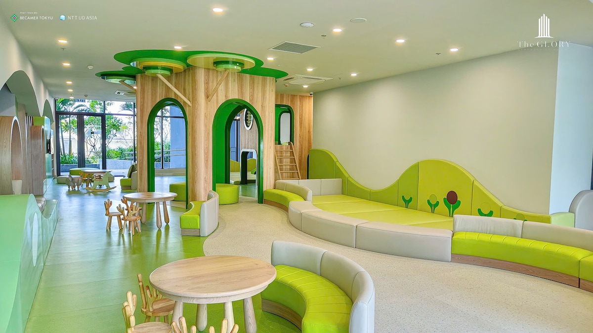 A modern play area features curved structures in various shades of green, complemented by light wooden tables and chairs. Soft seating areas are arranged along the walls, with large windows allowing natural light to fill the space. The overall design encourages a playful and inviting environment.