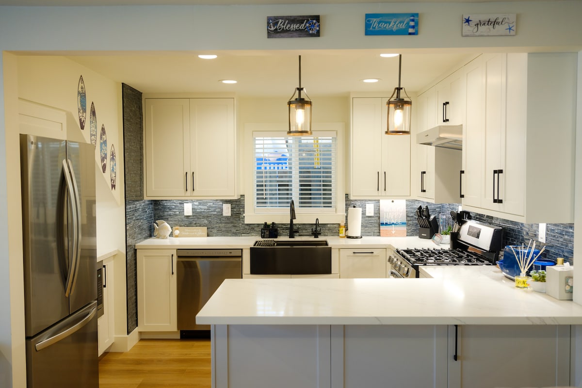 A modern kitchen is showcased with white cabinetry and stainless steel appliances. A spacious island provides additional counter space, complemented by decorative lighting above. Subtle blue tones are integrated into the backsplash, enhancing the inviting atmosphere. Natural light filters through the window, illuminating the area.