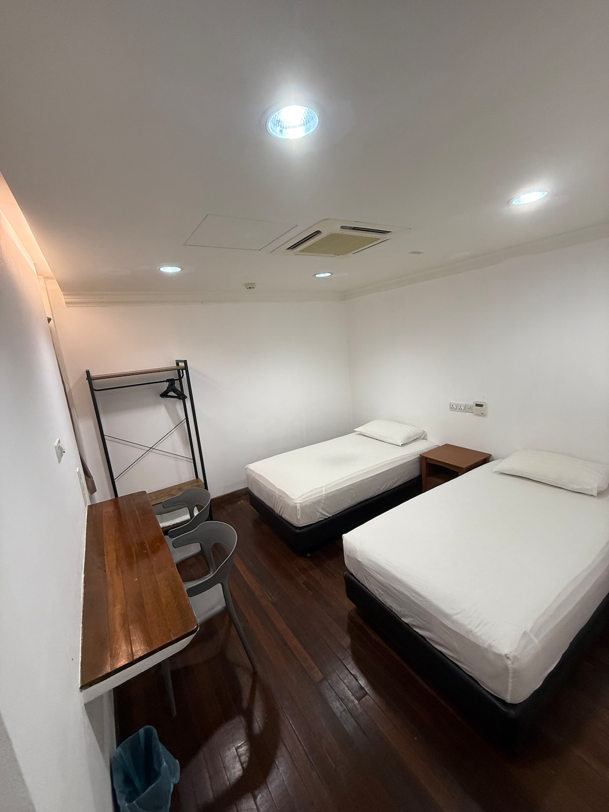 A well-lit room features two individual beds with white bedding, providing a clean and simple aesthetic. A wooden desk and chair create a functional workspace. A clothing rack is positioned against one wall, while the dark wood floor adds warmth to the overall design.