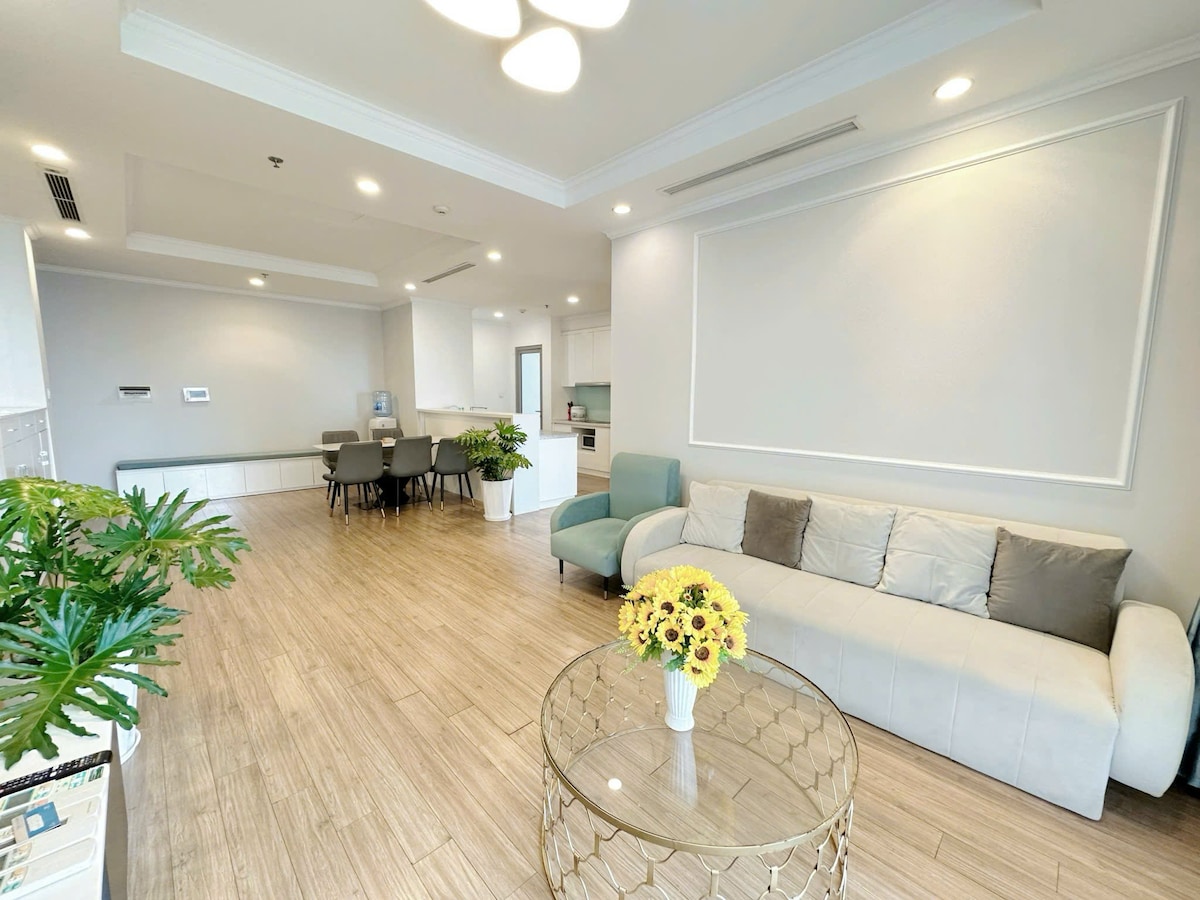 A well-lit living area features a light-colored sofa paired with decorative pillows. A glass coffee table sits at the center, adorned with a floral arrangement. Nearby, a dining table accommodates several chairs, while large windows allow natural light to enhance the space.