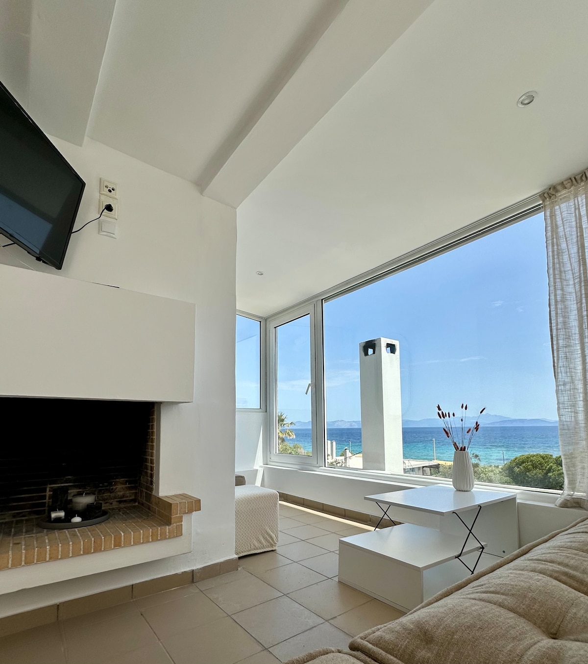 A bright living area is presented with large windows, offering views of the sea. A comfortable sofa is positioned near a modern fireplace, while a small side table adds functionality. Light colors and minimalistic decor create a relaxing environment infused with natural light.