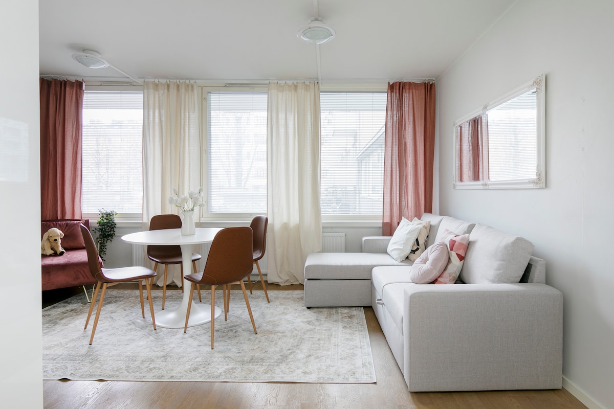 A bright living area features large windows draped with light curtains, allowing natural light to fill the space. A round dining table is surrounded by four wooden chairs, while a sectional sofa offers comfortable seating. A cozy rug is placed under the coffee table, completing the inviting atmosphere.