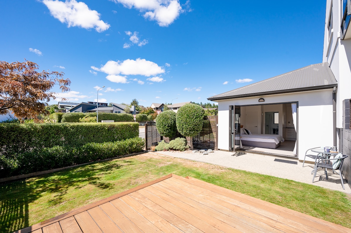 A sunny courtyard garden is showcased, featuring a wooden deck and well-maintained grass. The studio's entrance is visible with wide glass doors, allowing for easy access and scenic views of the surrounding greenery and blue sky.