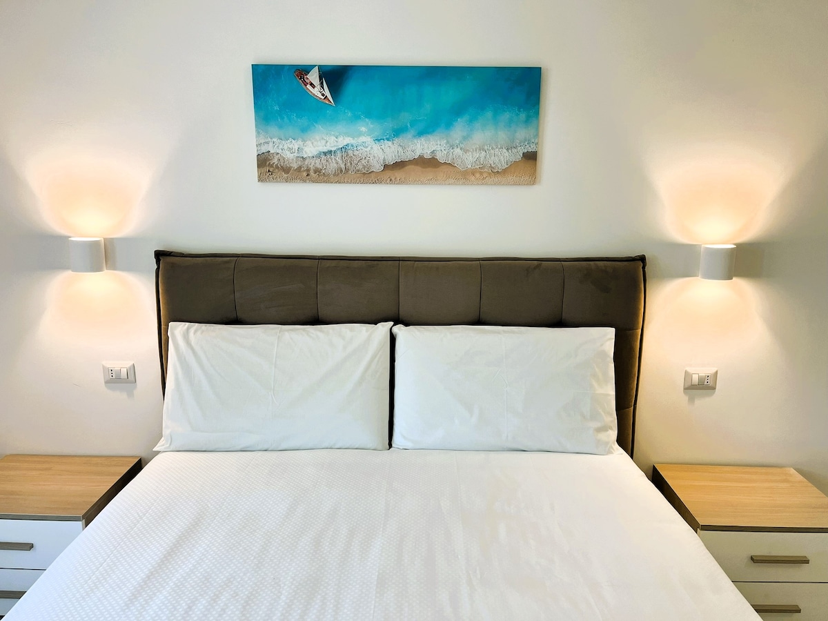 A spacious bed with crisp white linens is framed by two modern bedside tables. Soft lighting is provided by wall-mounted lamps on either side. Above the bed, a canvas print depicts a serene beach scene with gentle waves and a boat, enhancing the relaxing atmosphere.