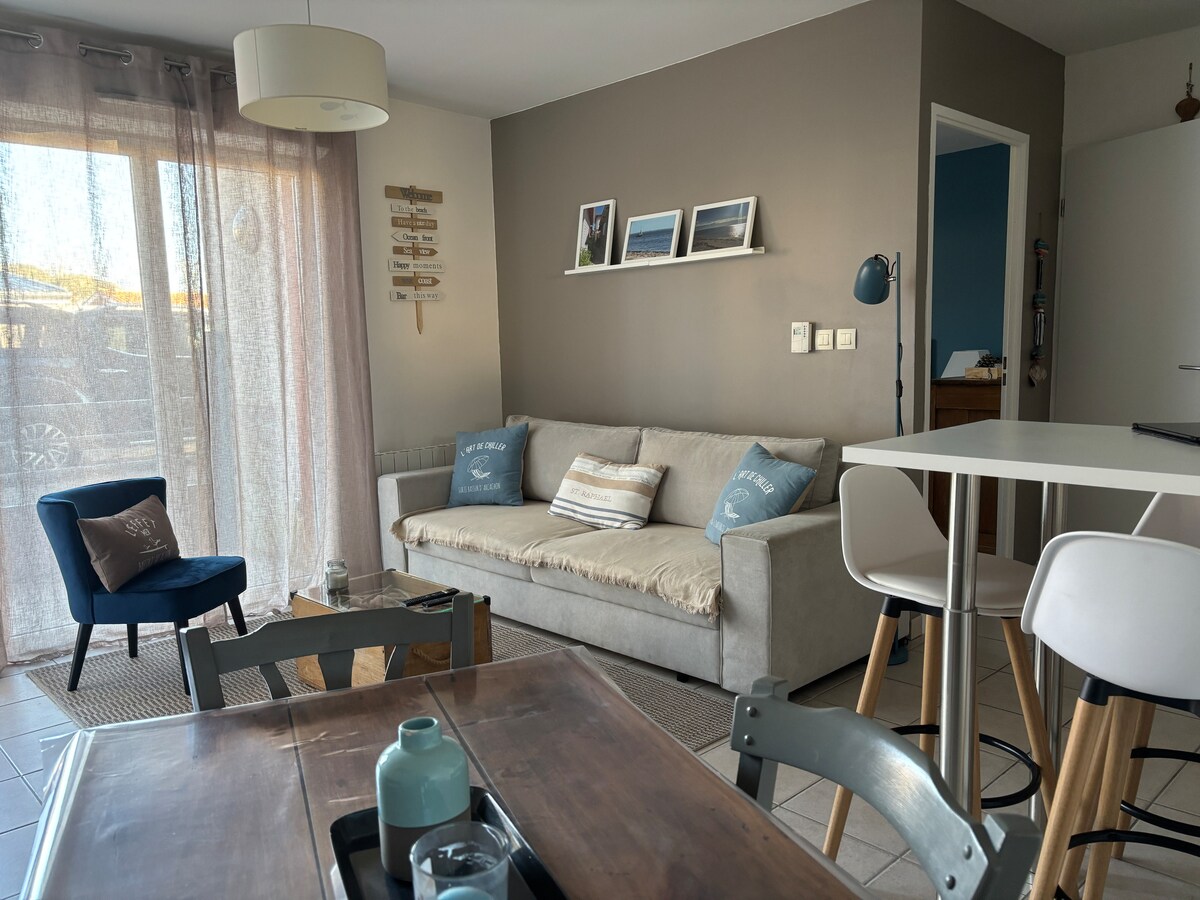 A living area is displayed with a light gray convertible sofa adorned with decorative pillows. A blue armchair and a wooden dining table with metal chairs are visible. Natural light filters through sheer curtains, illuminating the neutral tones of the decor.