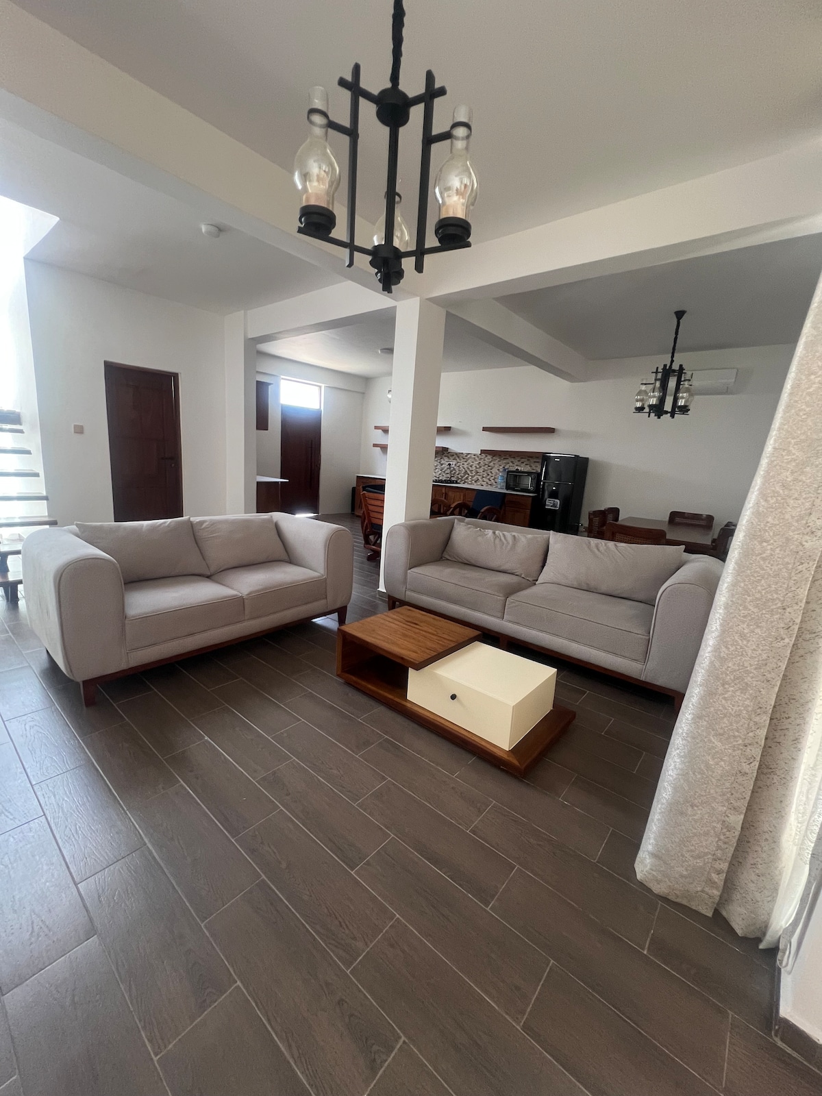 A bright living area features two light-colored sofas positioned around a wooden coffee table. A spacious kitchen area is visible in the background, complemented by wood furnishings. Natural light enters through windows, creating a cheerful atmosphere in the open space.