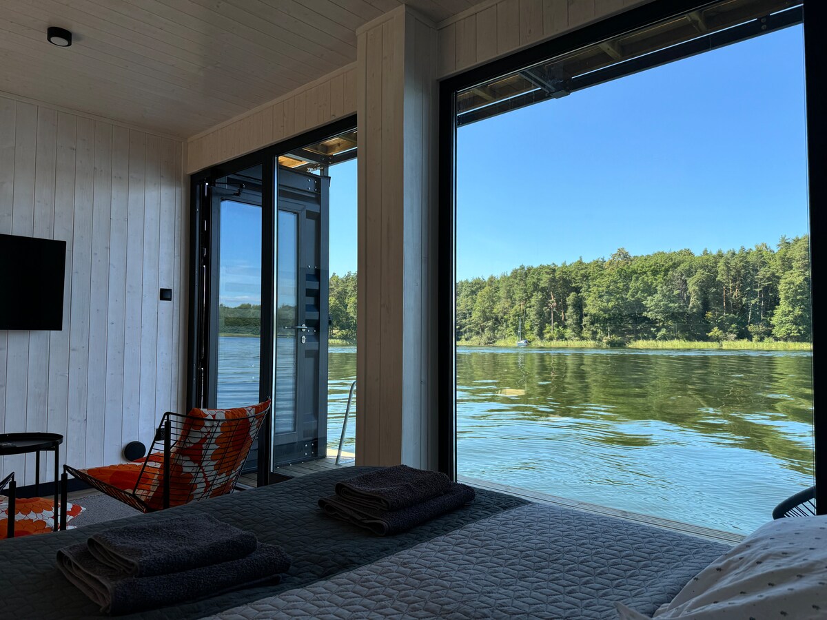 A serene interior space features large glass doors that open to a scenic view of the water and lush greenery. A cozy sleeping area is presented, with neatly arranged towels on the bed, enhancing the inviting atmosphere.
