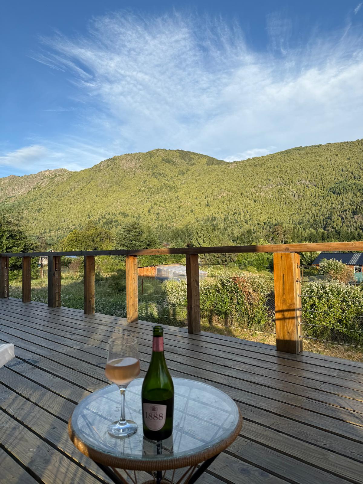 A wooden deck offers scenic views of rolling green mountains under a clear blue sky. A small round table is set with a glass of sparkling wine and an opened bottle, inviting relaxation in a tranquil outdoor space.
