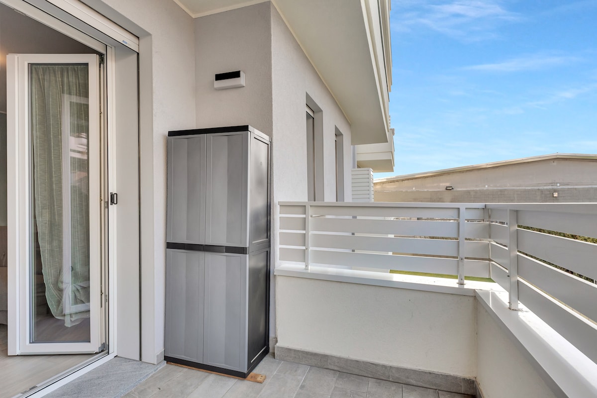 A private balcony is depicted, featuring a sleek grey storage unit against the wall. The space is surrounded by a white railing, providing an unobstructed view of the sky. Natural light enhances the clean and modern aesthetic of the area.