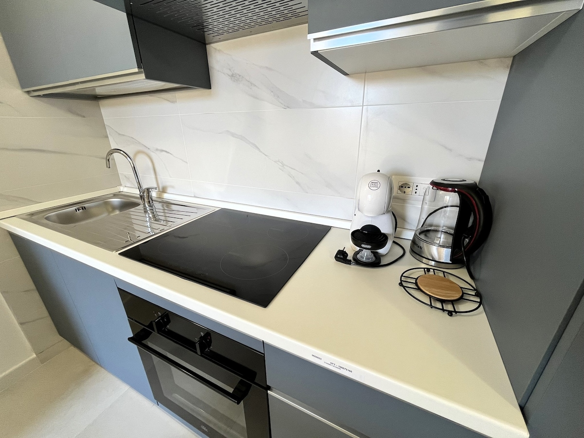 The kitchen features sleek cabinetry and a modern countertop. Equipped with an induction cooktop, oven, and stainless-steel sink, it also includes a coffee maker and kettle for convenience. Clean lines and neutral colors create a functional cooking area.
